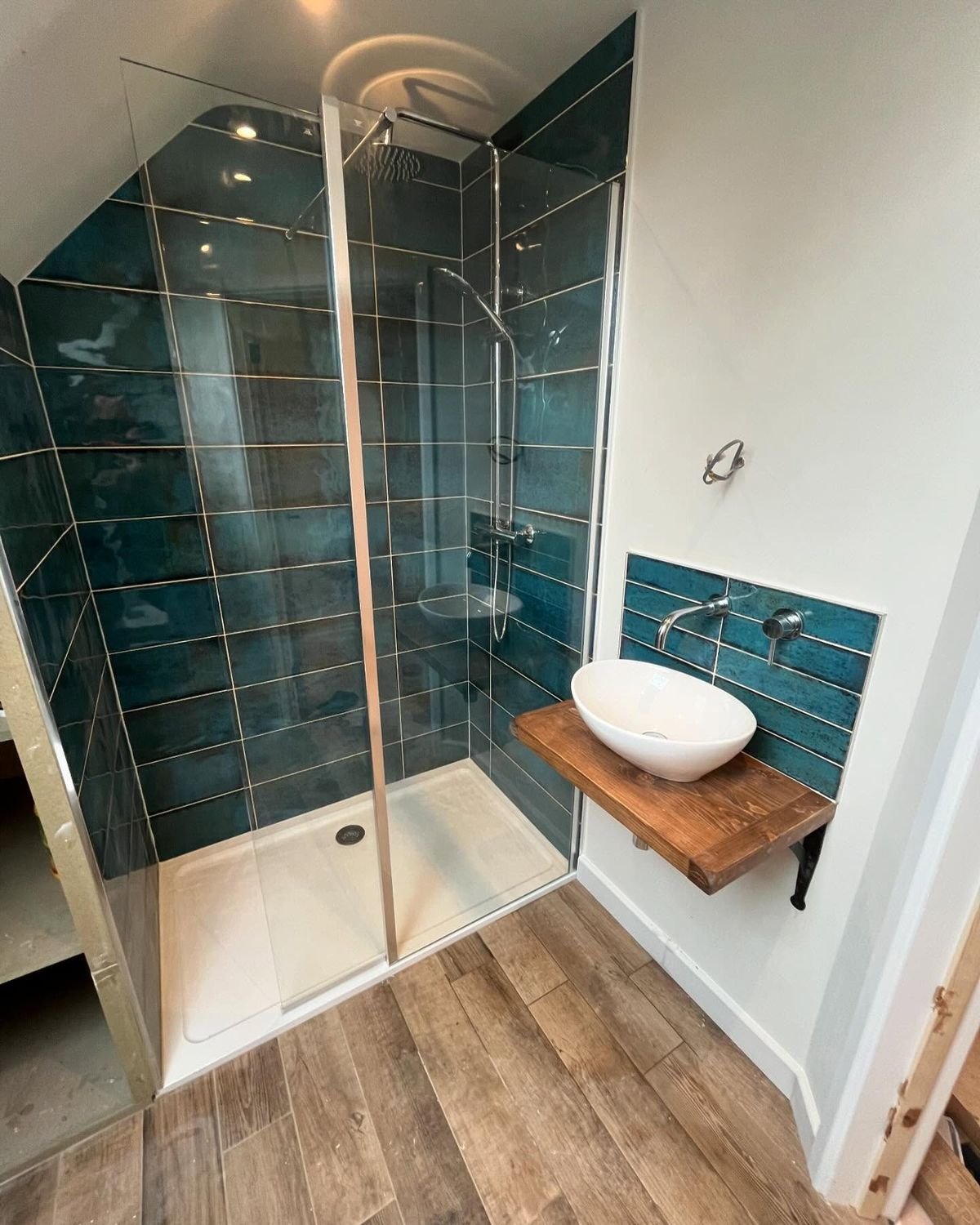 En suite shower with deep teal tiles and a floating oak shelf holding a basin